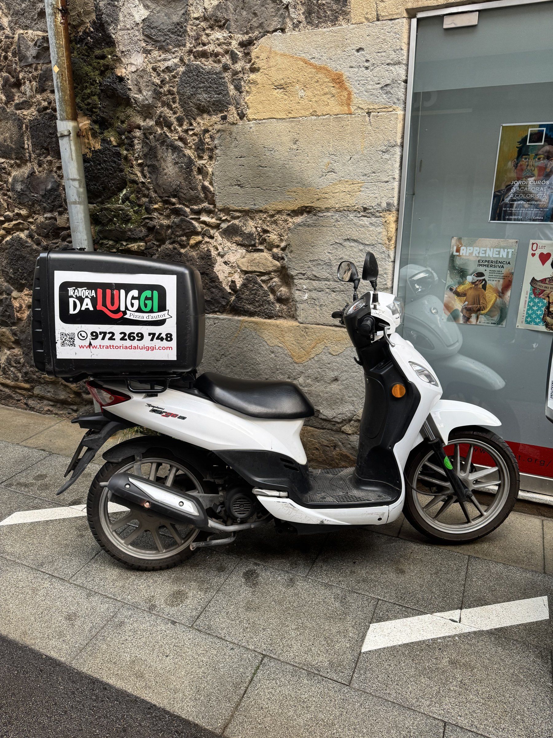 La moto de repartiment de Trattoria da Luiggi, amb el logotip i el telèfon al bagul, aparcada davant d'un mur de pedra