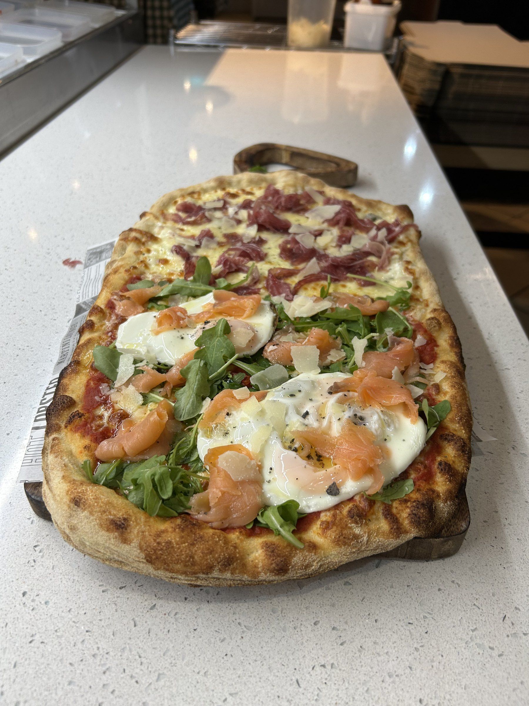 Pizza al taglio sobre una taula de fusta, meitat amb salmó, ou i ruca i meitat amb cecina i parmesà