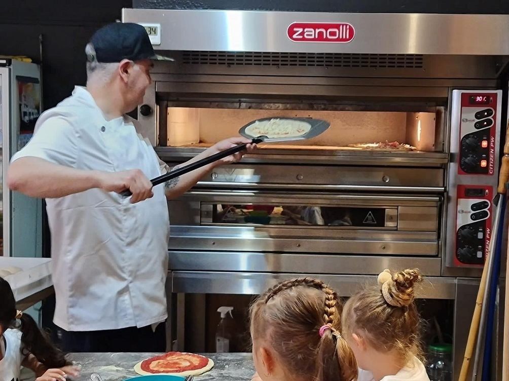 En Luiggi enforna una pizza durant el taller amb els nens de l'escola, que miren d'esquena
